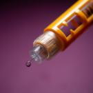 Close-up of an orange syringe with a droplet at the tip against a purple background.